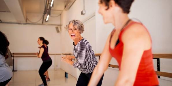 Zwei Frauen nehmen an einem Fitnesskurs teil und lachen dabei. Im Hintergrund sieht man weitere Teilnehmer.