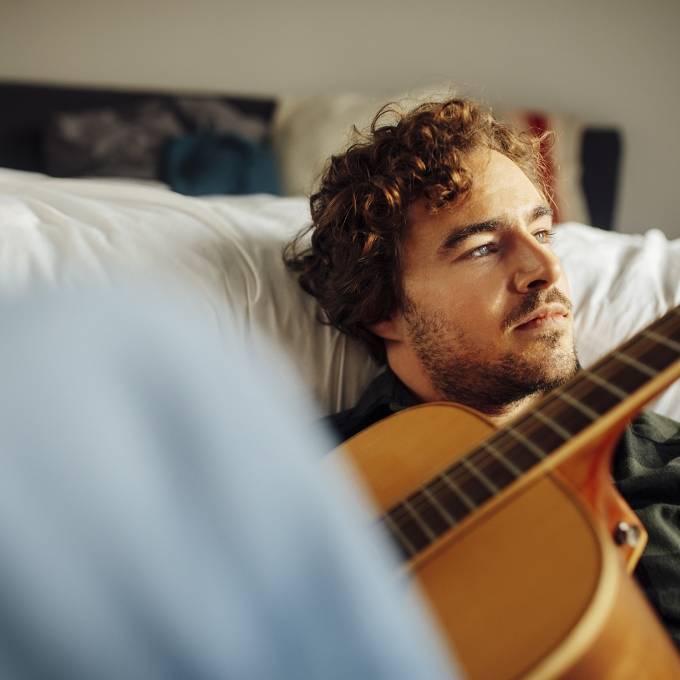 Mann sitzt auf dem Boden vor einem Bett und spielt Gitarre.