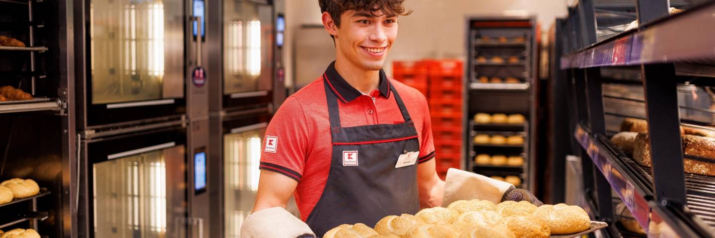 Junger Verkaufsmitarbeiter von Kaufland mit einem Blech frischgebackener Brötchen, die er gleich in die SB-Backwarenregale einsortiert.